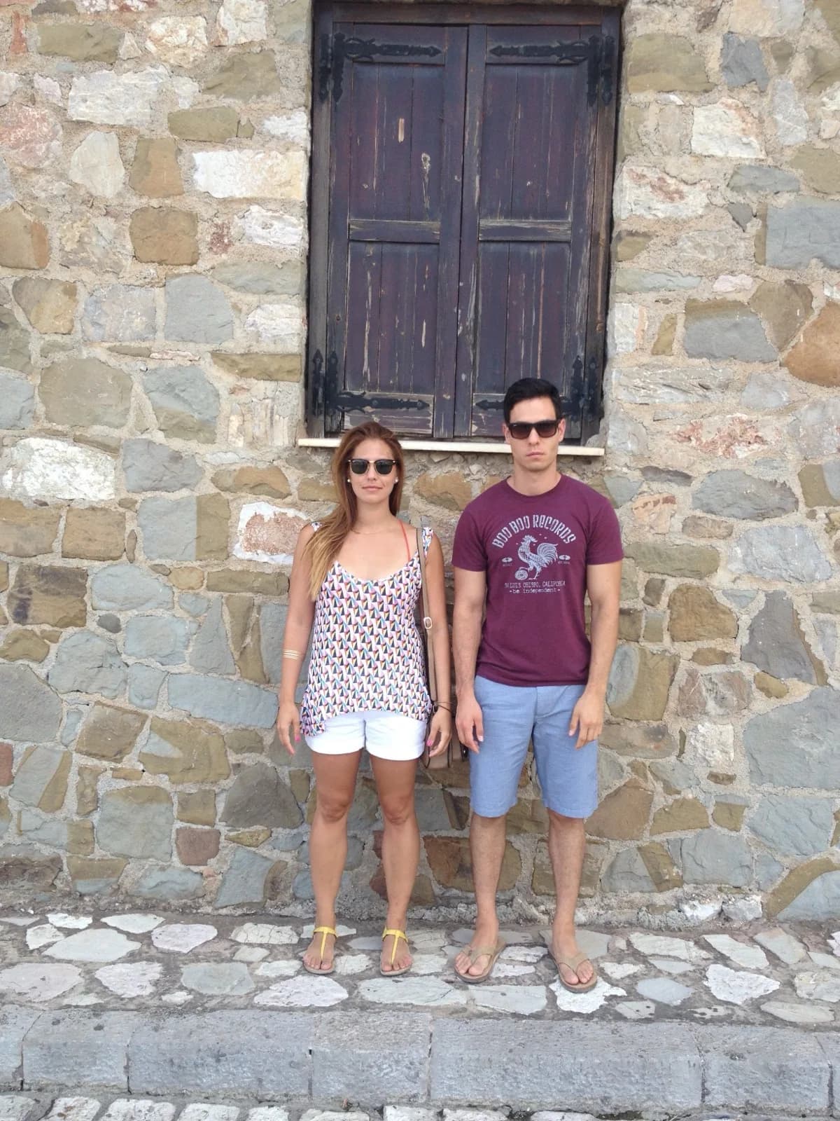 Amie and Adam Watanabe standing against a stone wall beneath a weathered wooden shutter while travelling