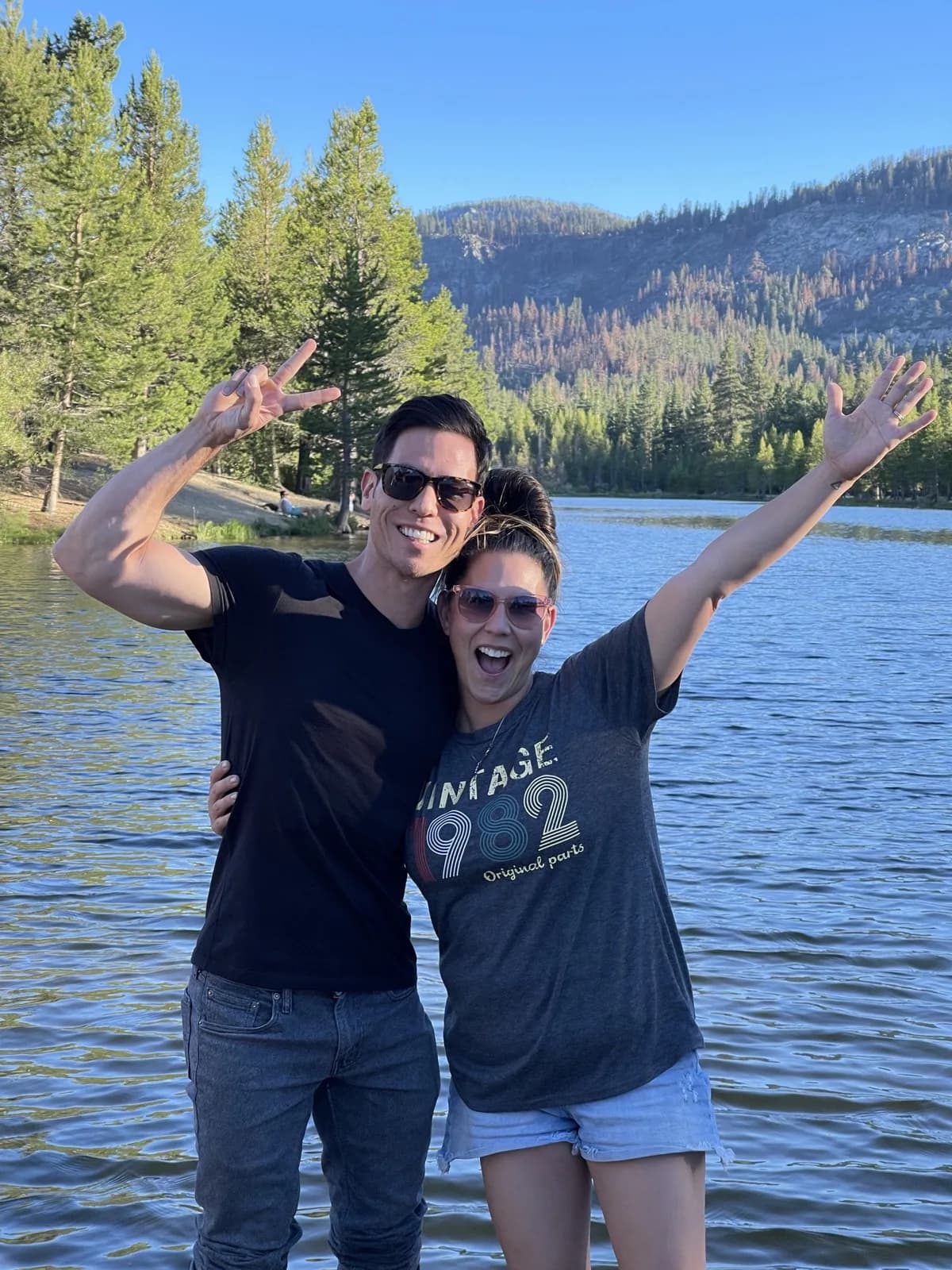 Adam and Amie Watanabe with their arms raised, laughing in front of a mountain lake in Northern California