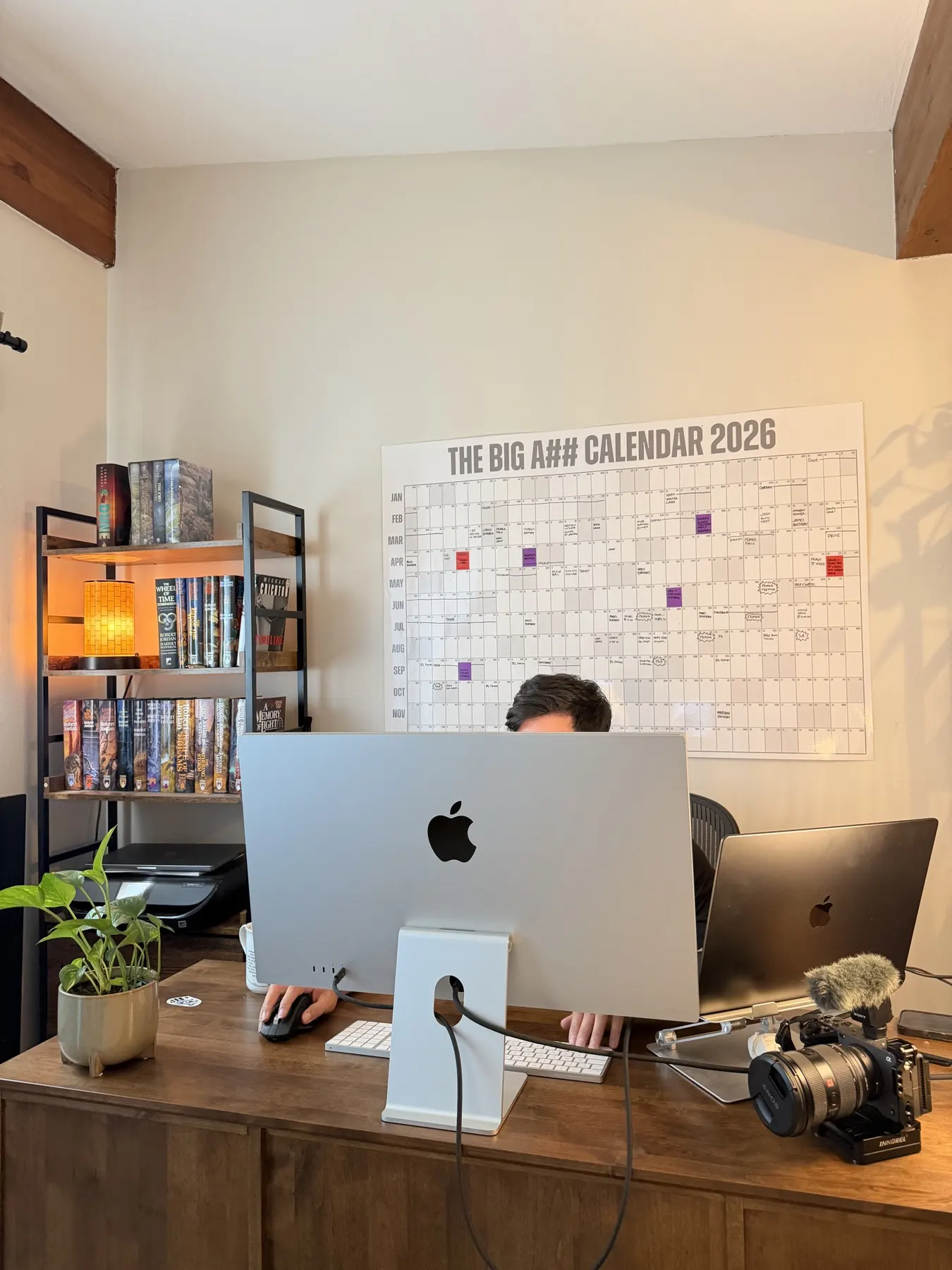 Adam Watanabe at his desk behind a large monitor, editing, with a wall planner and camera gear around him