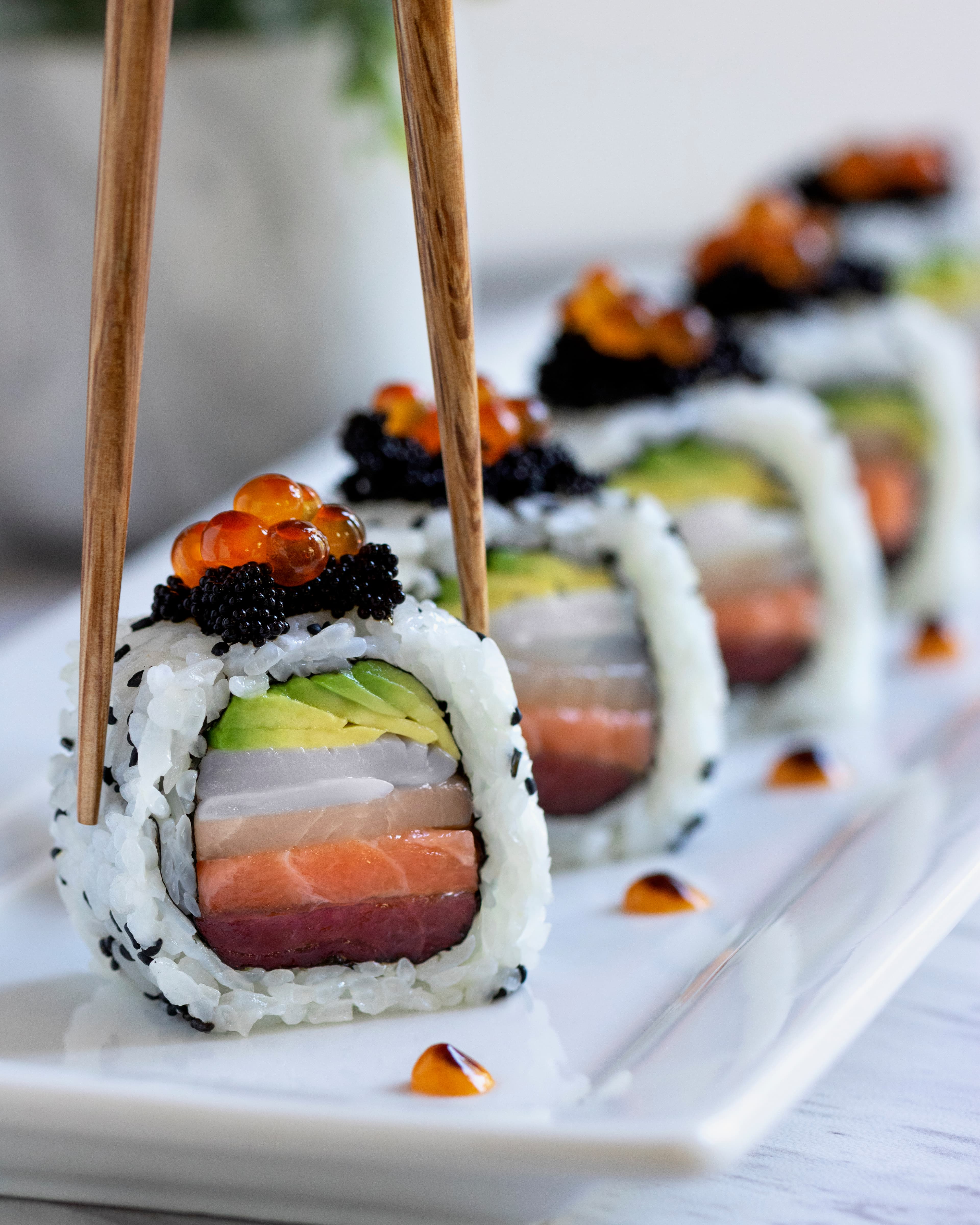 Row of sesame-crusted sushi rolls layered with avocado, yellowtail, tuna, and salmon, topped with roe and caviar