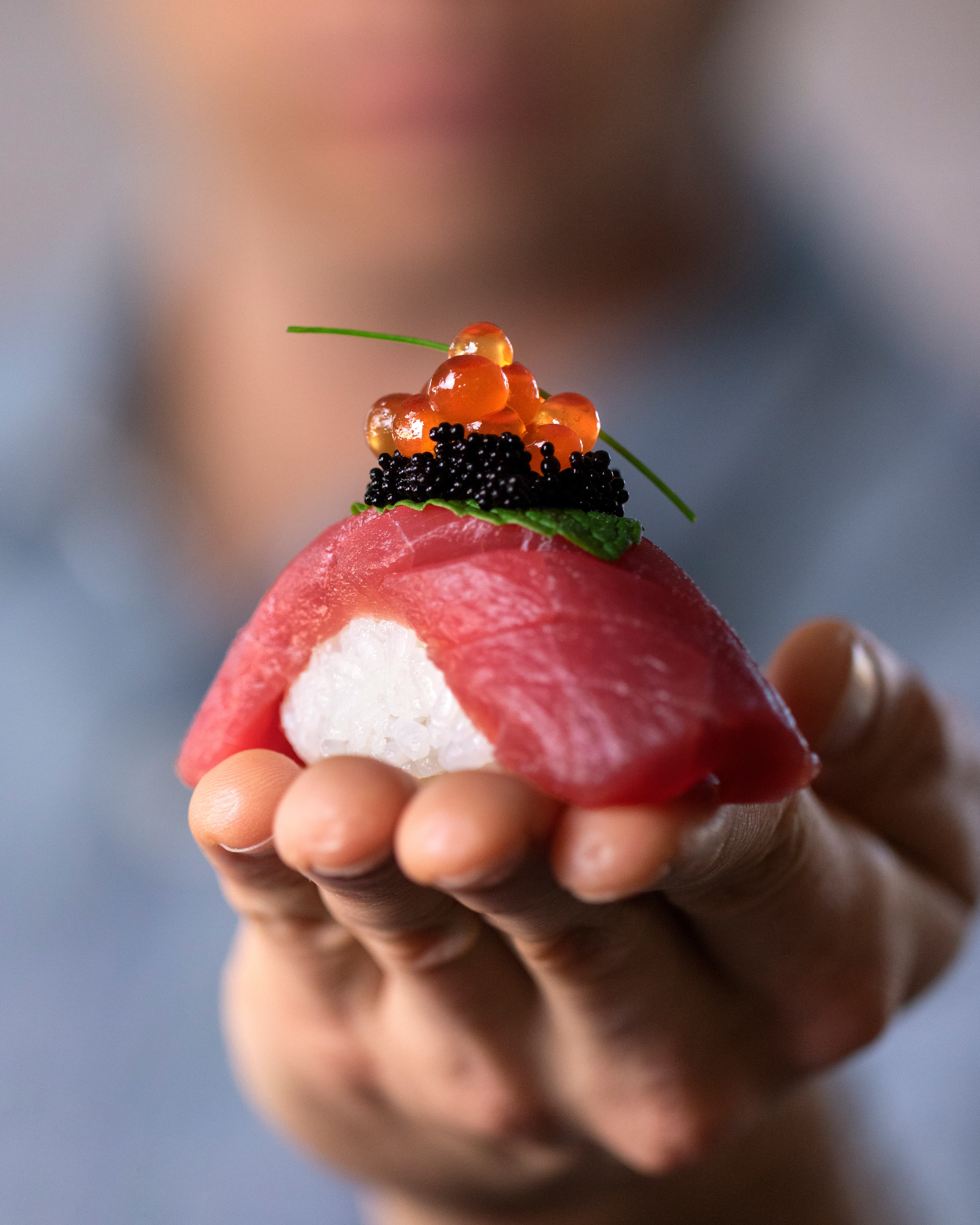 Hand holding a tuna nigiri topped with salmon roe, caviar, and a shiso leaf