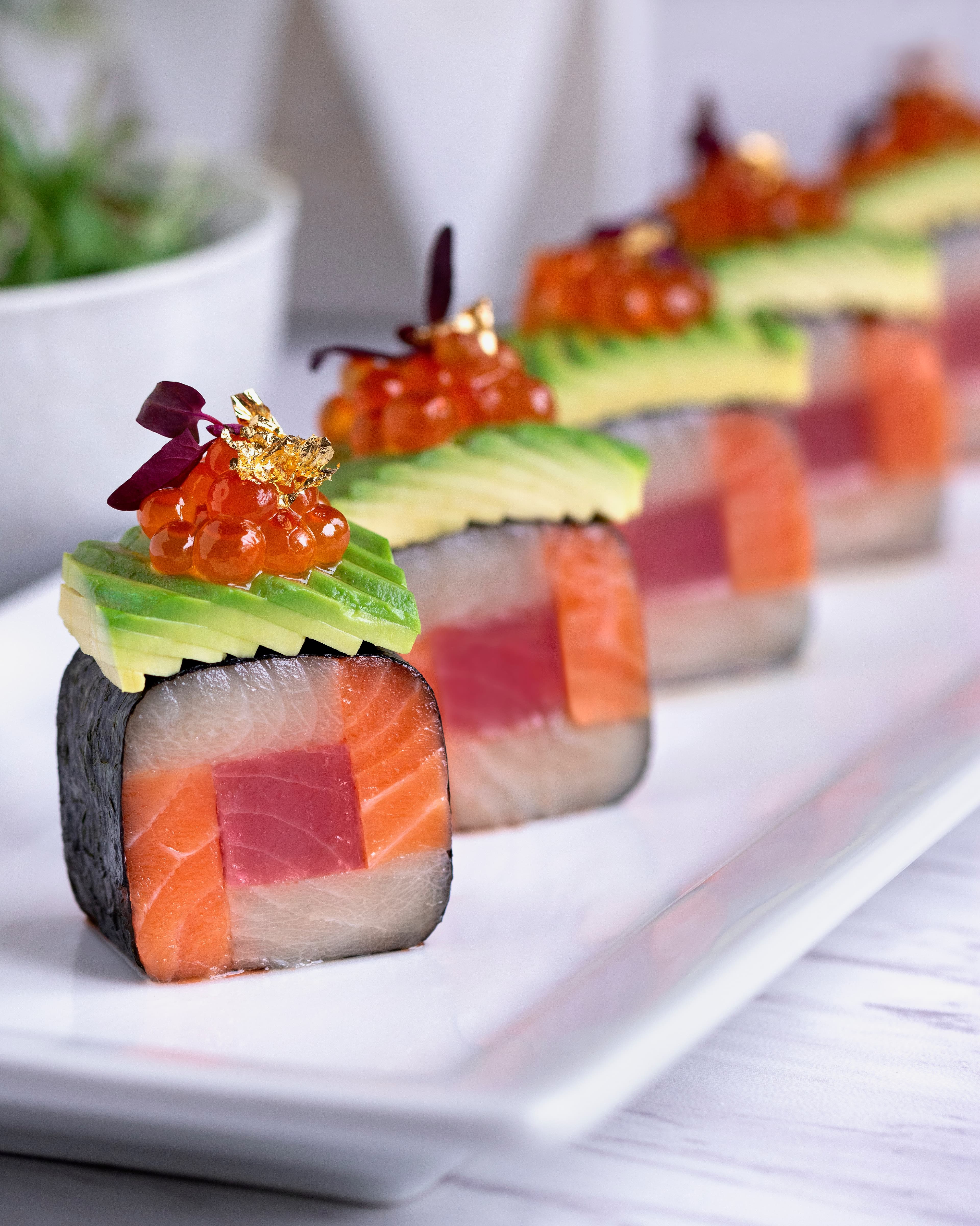 Row of sushi rolls layered with tuna, salmon, and yellowtail, topped with avocado, salmon roe, and gold leaf