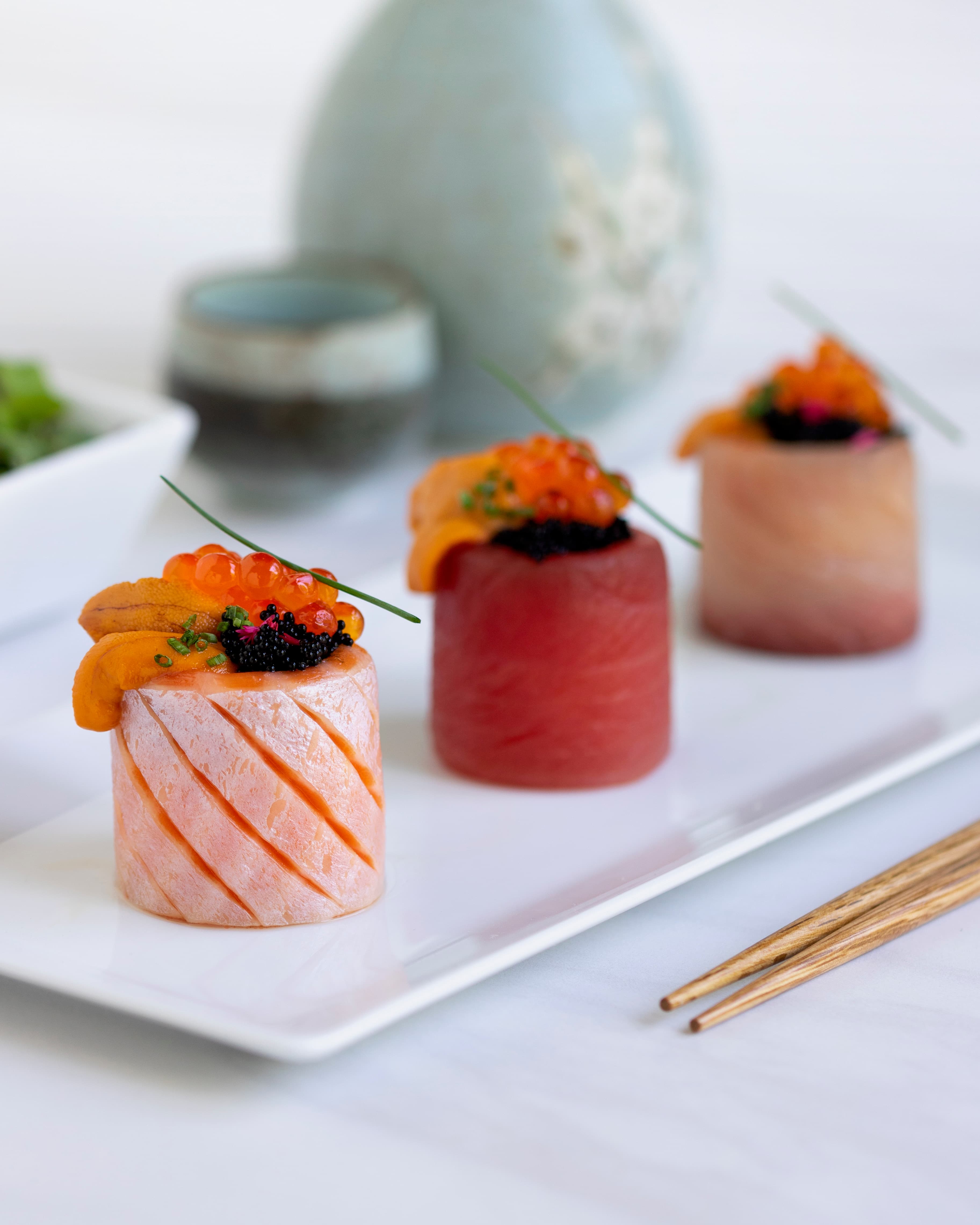 Three cylindrical sushi pieces of salmon, tuna, and yellowtail topped with sea urchin, roe, and caviar, served with sake cups