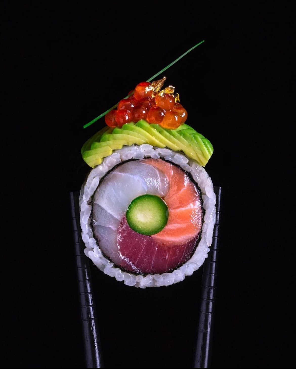 Sushi roll topped with avocado, salmon roe, and gold leaf, filled with yellowtail, salmon, and tuna, held between chopsticks